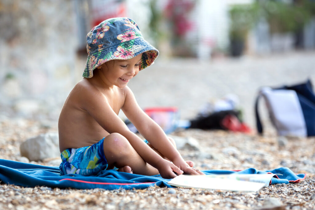 Cute blond toddler boy, reading a book on the beach, sitting on his towel