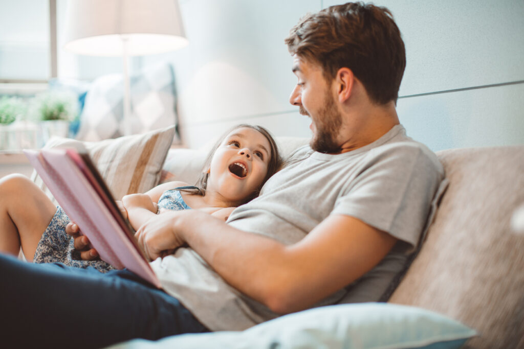 Father and daughter enjoying at home. Sitting on bed and reading book together.